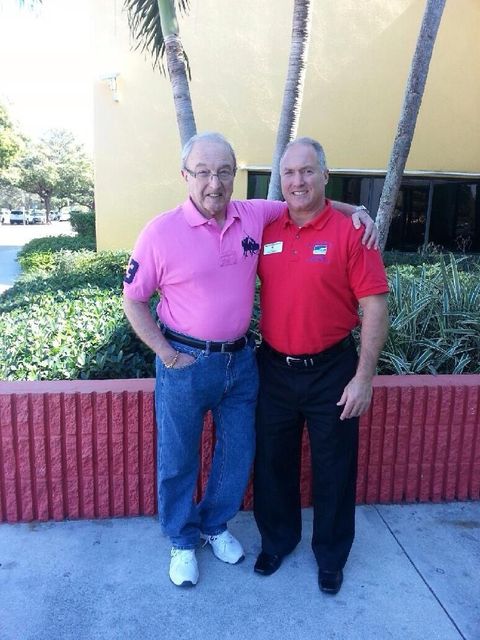 Two men standing and smiling outside a building, one wearing a pink shirt and the other in a red shirt.