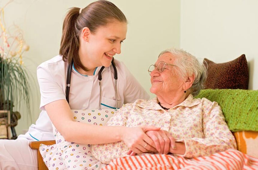 A caregiver smiling at an elderly woman while holding her hand in a comforting manner.